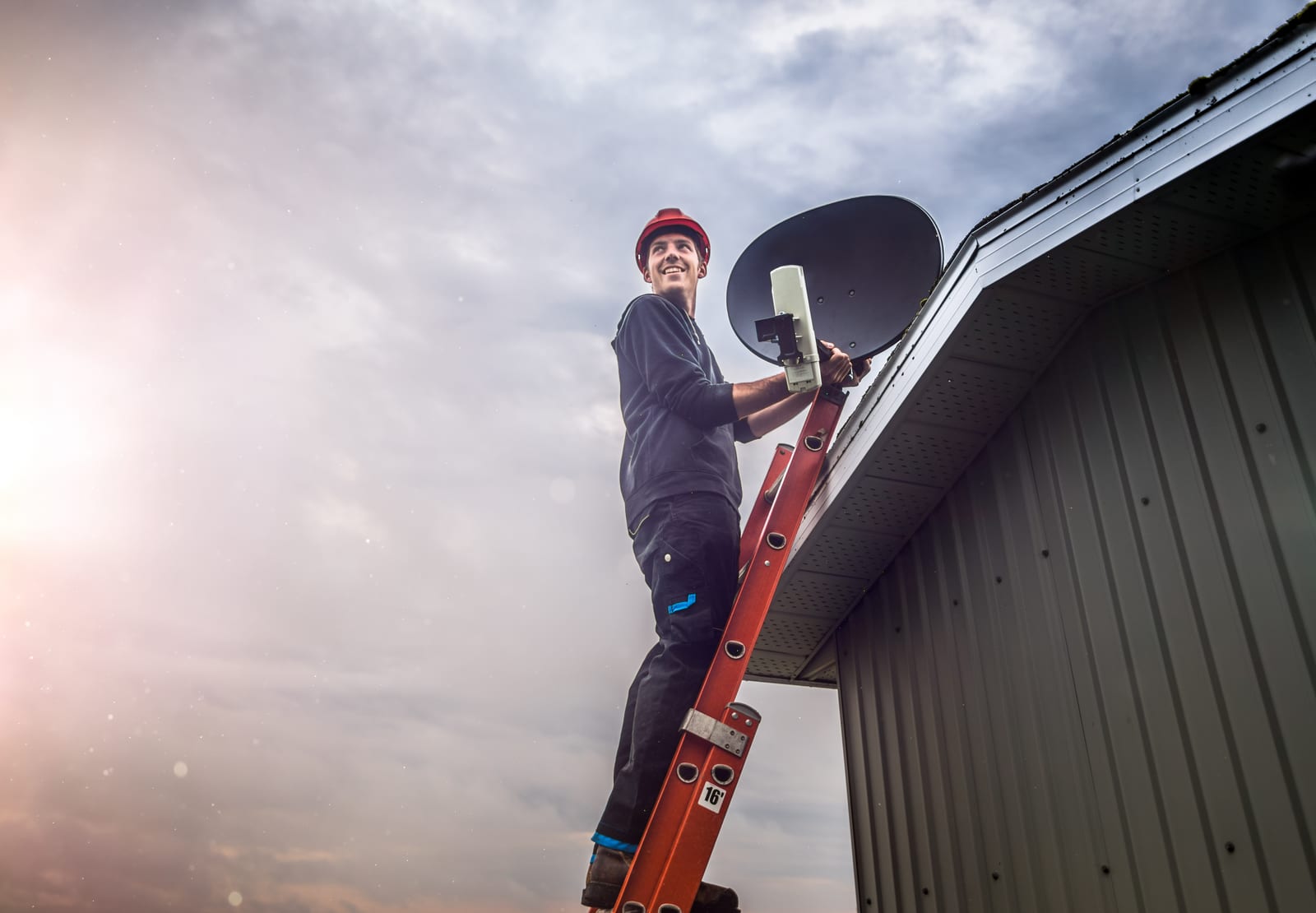 A technician up a ladder fitting an antenna to the side of a building